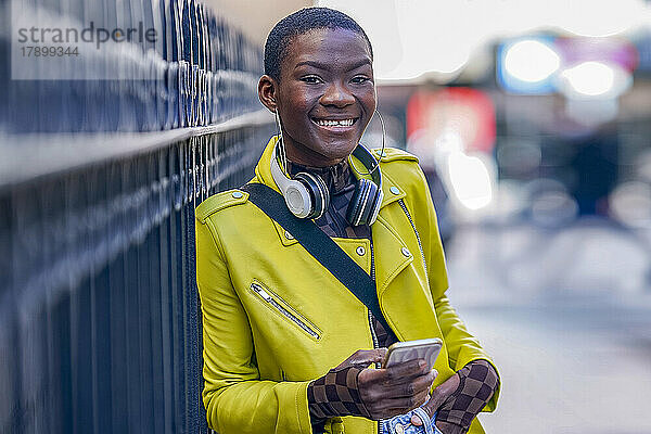Lächelnde Frau mit Smartphone lehnt am Zaun