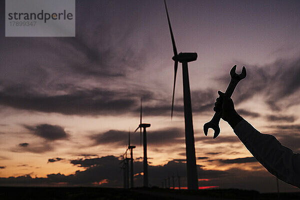 Silhouette eines Ingenieurs  der bei Sonnenuntergang einen Schraubenschlüssel neben Windkraftanlagen im Windpark hält