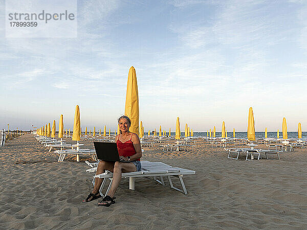 Glückliche ältere Geschäftsfrau sitzt mit Laptop am Strand