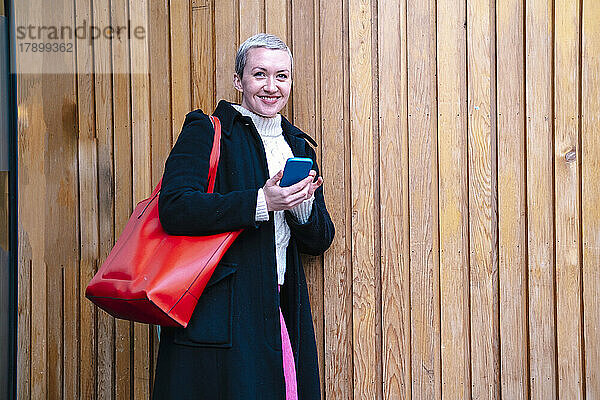 Glückliche Frau mit Mobiltelefon  das an einer Holzwand lehnt