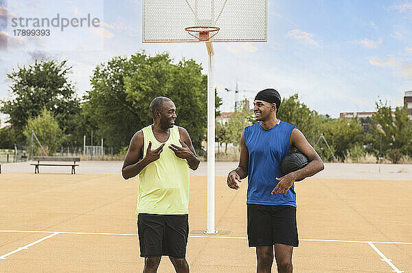 Lächelnder junger Mann hält Ball und unterhält sich mit Vater auf dem Basketballplatz