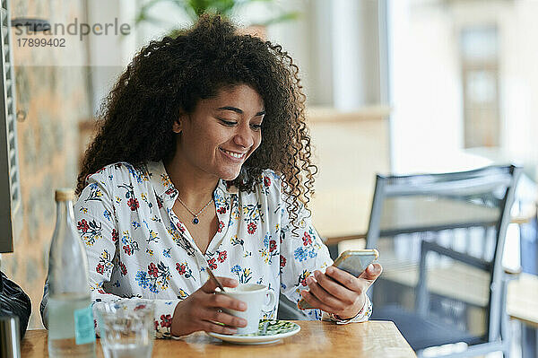 Glückliche junge Frau sitzt mit Kaffeetasse und Smartphone im Café
