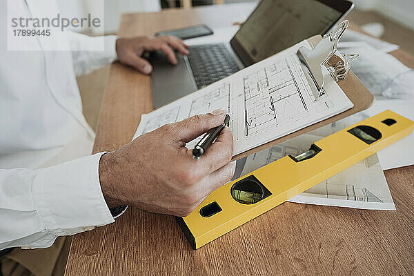 Hände des Architekten halten einen Stift in der Hand und zeigen auf einen Bauplan auf dem Klemmbrett im Büro