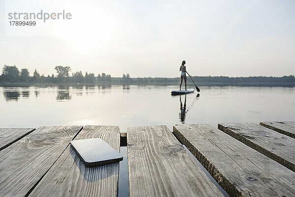 Mobiltelefon am Steg mit Frau beim Standup-Paddleboarding im Hintergrund bei Sonnenuntergang