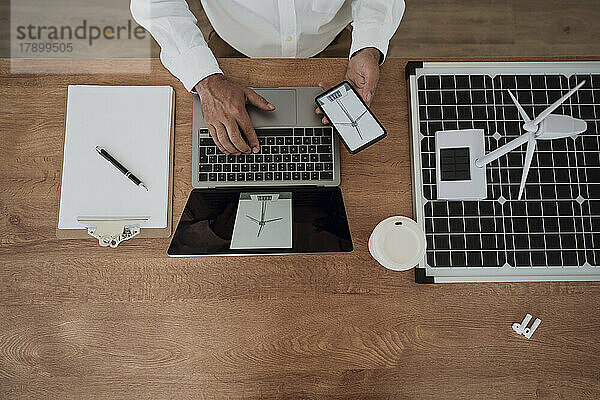 Hände eines Architekten mit Smartphone und Laptop  der neben einer Windkraftanlage auf einem Solarpanel am Schreibtisch sitzt