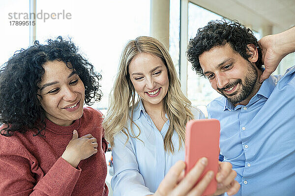Lächelnde blonde Geschäftsfrau  die im Büro ein Selfie mit Kollegen auf dem Smartphone macht