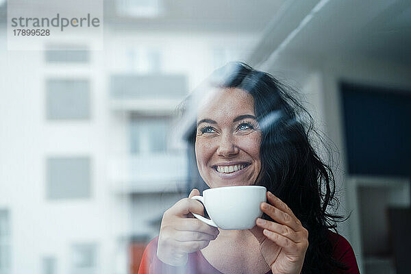 Glückliche Frau hält Kaffeetasse durch Glas gesehen