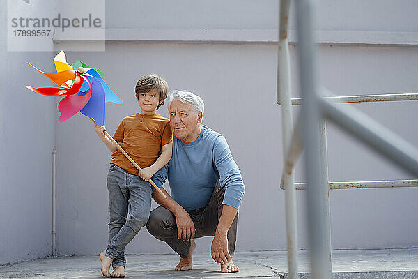 Nachdenklicher älterer Mann neben seinem Enkel  der ein buntes Windradspielzeug im Flur hält