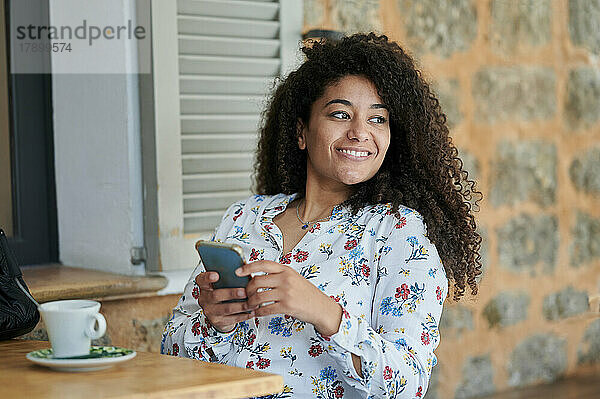 Nachdenklich lächelnde junge Frau sitzt mit Mobiltelefon im Café