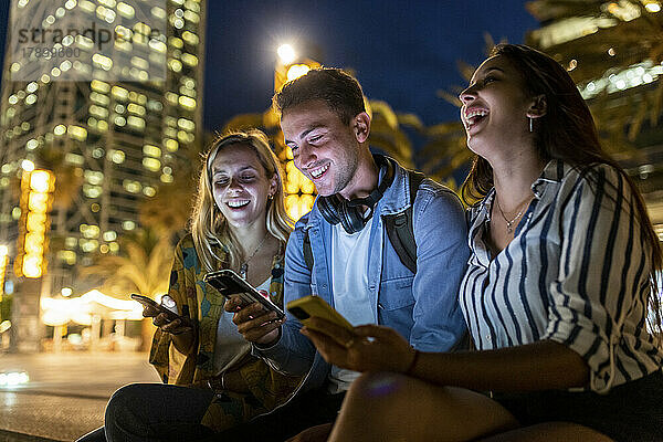 Cheerful women sitting by friend using smart phone at night
