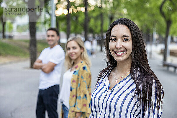 Lächelnde junge Frau mit langen Haaren steht vor Freunden im Park