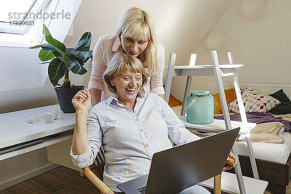 Blonde Frau mit Mutter sitzt mit Laptop auf Stuhl im Schlafzimmer