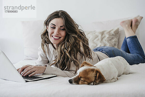 Lächelnde Frau schaut Hund an  der mit Laptop im Bett liegt