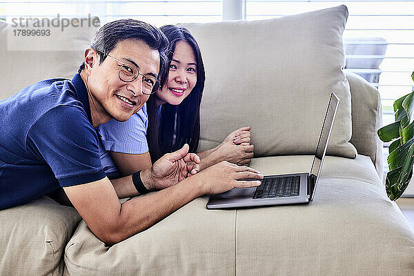 Happy mature couple with laptop lying on sofa at home