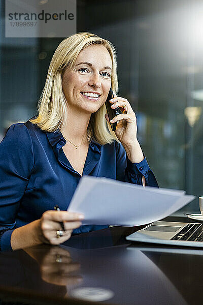 Lächelnde Geschäftsfrau mit Dokumenten  die am Tisch im Café auf dem Smartphone spricht