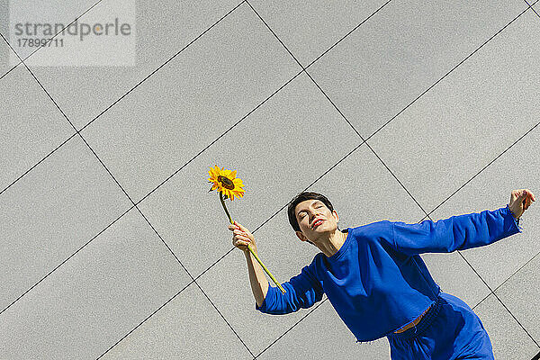 Frau mit geschlossenen Augen hält an einem sonnigen Tag eine Sonnenblume vor der Wand