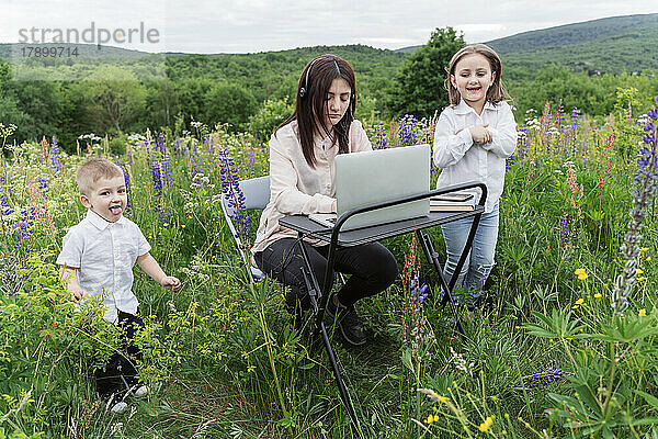 Verspielte Kinder von Mutter  die auf der Wiese am Laptop arbeitet