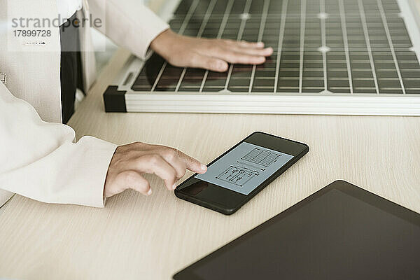 Nahaufnahme einer Frau  die ihr Mobiltelefon mit Solarpanel auf dem Schreibtisch benutzt