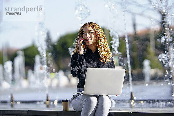 Glückliche Geschäftsfrau  die vor dem Brunnen sitzt und mit ihrem Smartphone spricht