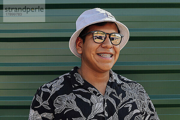 Glücklicher junger Mann mit Sonnenbrille und Hut vor der Wand