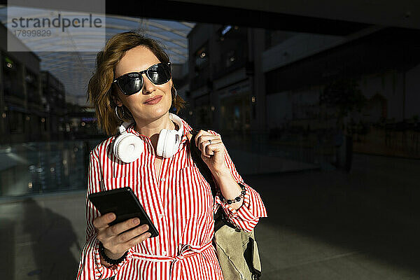 Lächelnde Frau mit Sonnenbrille und Mobiltelefon im Einkaufszentrum