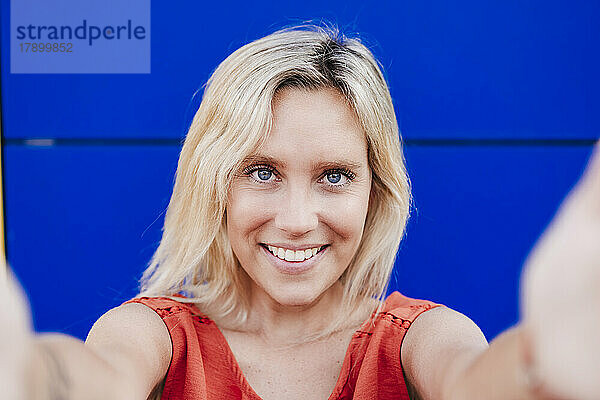 Happy young woman with blond hair taking selfie in front of blue wall