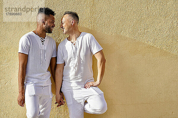 Cheerful gay couple holding hands in front of wall on sunny day
