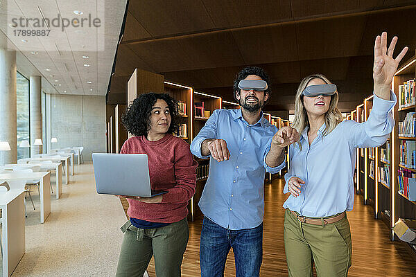 Mann und Frau mit VR-Brille gestikulieren auf einen Kollegen  der in der Bibliothek einen Laptop hält