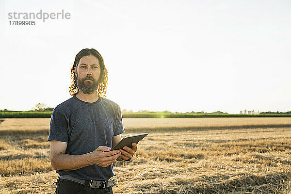 Bärtiger Bauer mit Tablet-PC steht an einem sonnigen Tag auf dem Bauernhof