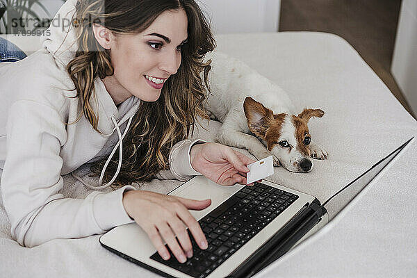 Junge Frau mit Kreditkarte erledigt Online-Einkäufe per Laptop und liegt zu Hause beim Hund
