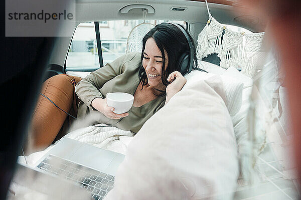 Lächelnde Frau mit Kopfhörern  die sich einen Film auf dem Laptop ansieht und im Kofferraum eines Autos Kaffee trinkt