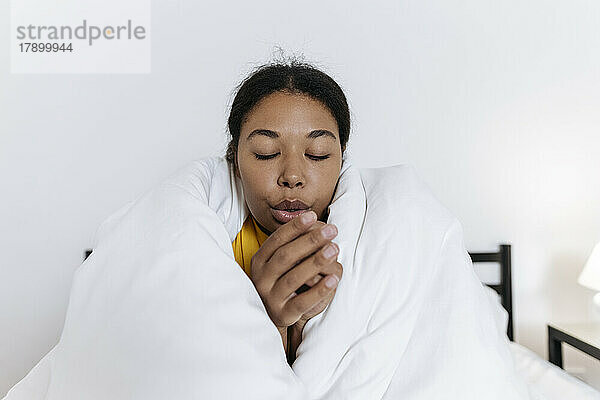 Frierende junge Frau sitzt in eine Decke gehüllt auf dem Bett und reibt sich die Hände