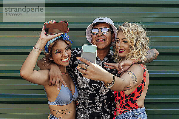 Friends having fun taking selfie through smart phone in front of green wall