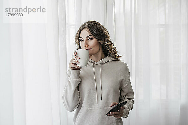 Frau mit Smartphone trinkt Kaffee vor weißem Vorhang zu Hause
