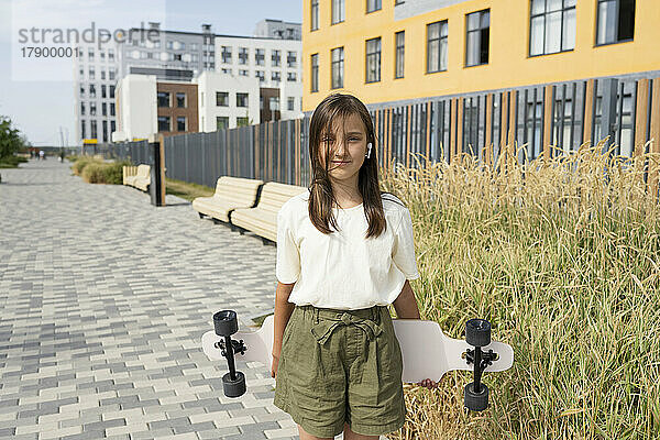 Lächelndes Mädchen steht mit Skateboard auf Fußweg