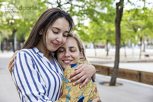 Lächelnde junge Frauen mit geschlossenen Augen umarmen sich im Park