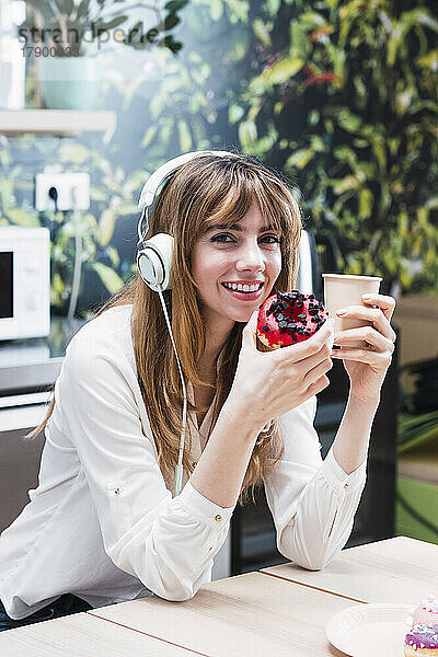Lächelnde Geschäftsfrau mit Kopfhörern sitzt mit Einwegkaffeetasse und Donut in der Cafeteria