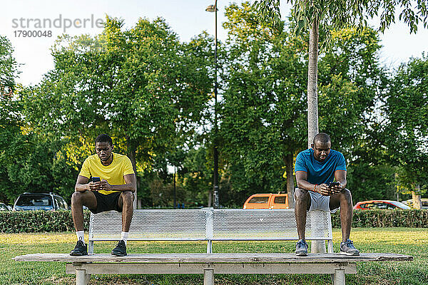 Young men using smart phones sitting on bench at park