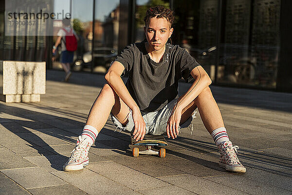 Selbstbewusste nicht-binäre Person sitzt auf einem Skateboard am Fußweg