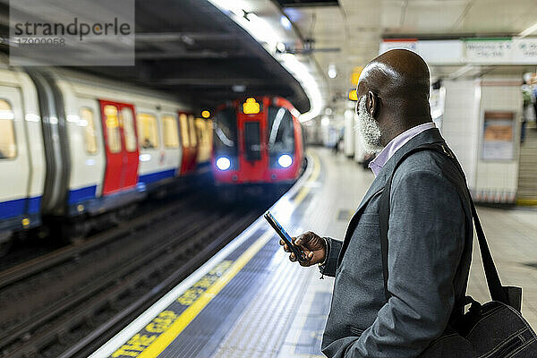Geschäftsmann schaut mit Smartphone auf U-Bahn  die am Bahnhof ankommt