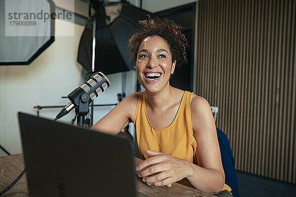 Reifer Radiomoderator lacht mit Mikrofon und Laptop auf dem Tisch im Studio