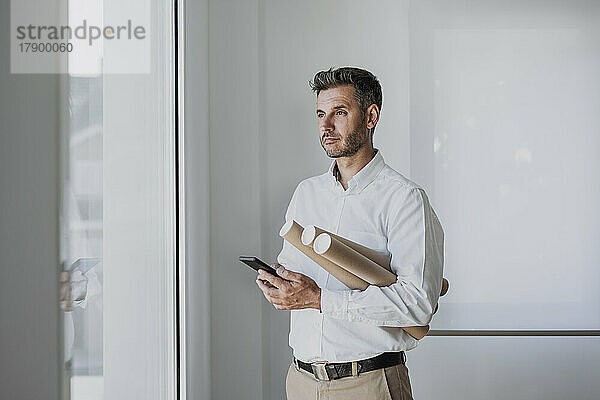 Nachdenklicher Architekt hält Mobiltelefon und aufgerolltes Papier in der Hand und schaut durch das Fenster im Büro