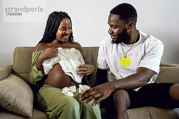 Schwangere Frau zeigt einem Mann  der zu Hause auf dem Sofa sitzt  Babykleidung