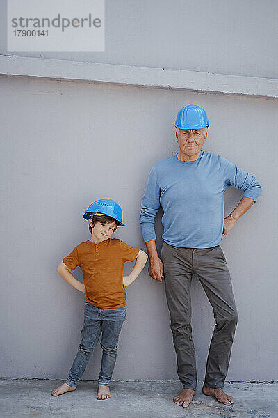 Selbstbewusster älterer Mann und Enkel mit Schutzhelmen stehen mit den Händen auf der Hüfte vor der Wand