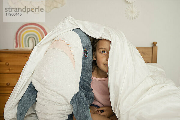 Mädchen versteckt sich unter Decke mit Haispielzeug im Kinderzimmer