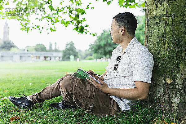 Junger Mann mit Sudoku-Buch lehnt an Baumstamm im Park