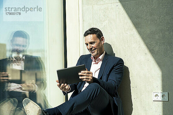 Lächelnder Geschäftsmann  der über einen Tablet-PC am Fenster im Büro Videoanrufe tätigt