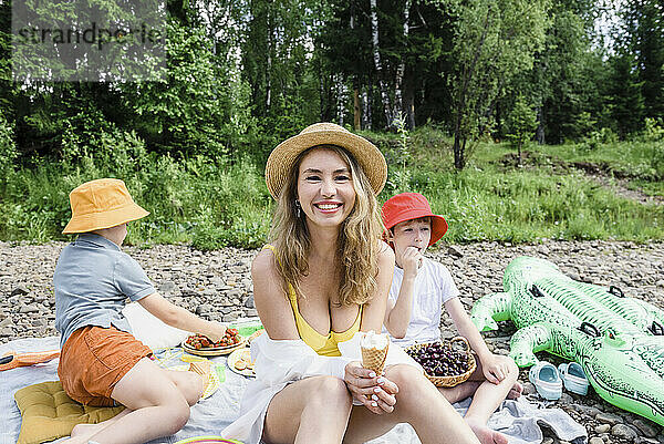 Lächelnde Frau hält Eis und sitzt mit Söhnen beim Picknick