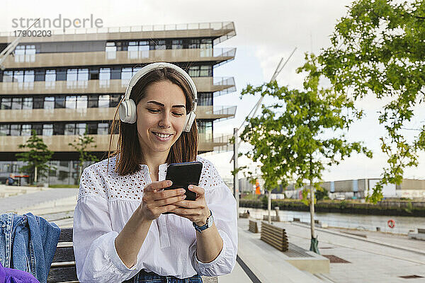 Glückliche Frau  die Musik über kabellose Kopfhörer hört und ihr Smartphone auf der Bank benutzt