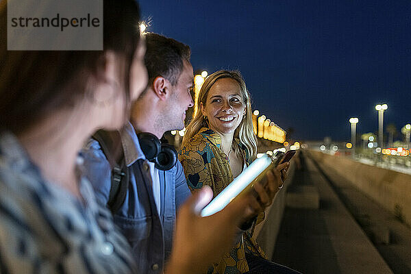 Glückliche Frau  die nachts mit Freunden spricht  die Smartphones in der Hand halten